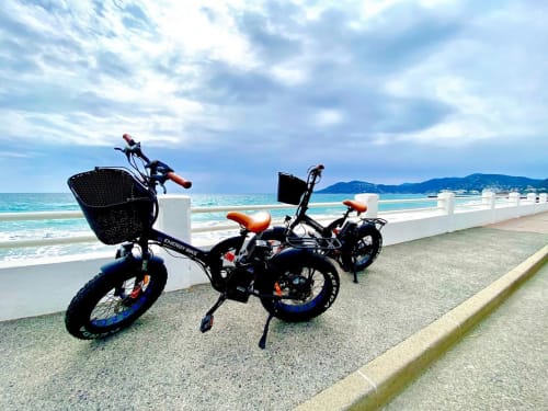 Deux vélos électriques avec paniers garés sur une promenade en bord de mer offrant vue sur les eaux turquoise et la plage de sable fin à Mandelieu-la-Napoule