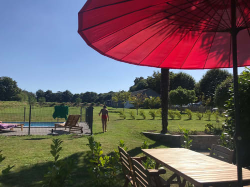 Parasol rouge ombrageant la terrasse en bois des Gîtes De L'odet à Clohars-Fouesnant, avec piscine et champs verdoyants