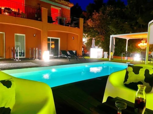 Vue nocturne de la piscine Villa Colina avec eau turquoise, bâtiment orange et mobilier jaune fluo à Mandelieu-la-Napoule