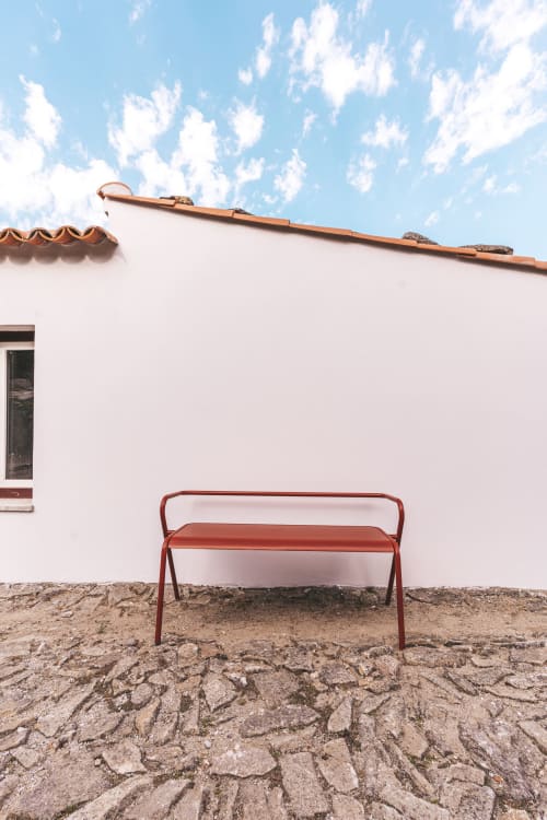Detalhe minimalista nos Moinhos d’Antoninha: banco de metal vermelho sobre um chão de pedra, junto a uma parede branca e um telhado tradicional, num ambiente que respira calma e autenticidade.