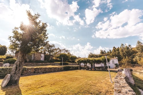 Área exterior dos Moinhos d’Antoninha: relva verde, árvores, casas de pedra branca e uma pérgula com mesas, sob um céu azul com nuvens esparsas. Um refúgio de tranquilidade e beleza natural.