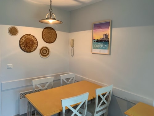 Salle à manger de l'appartement Belle Île en Mer avec table jaune, chaises blanches, paniers tressés muraux et affiche côtière