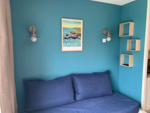 Chambre turquoise lumineuse avec coin salon bleu, œuvres côtières, étagères en bois et appliques murales à l'appartement Locmaria