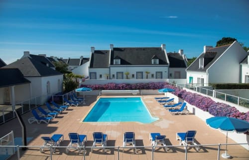 Piscine du resort Loc'Maria Belle Île en Mer avec transats bleus, fleurs violettes et immeubles blancs sous ciel bleu