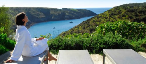 Femme savourant un repas face à la baie turquoise de Locmaria, Belle Île en Mer, avec littoral luxuriant et bateaux