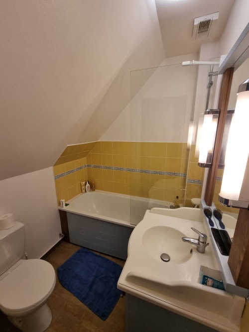 Salle de bains moderne aux murs en carrelage jaune, lavabo blanc, toilettes et baignoire avec paroi vitrée aux Locations Loc'Maria, Belle