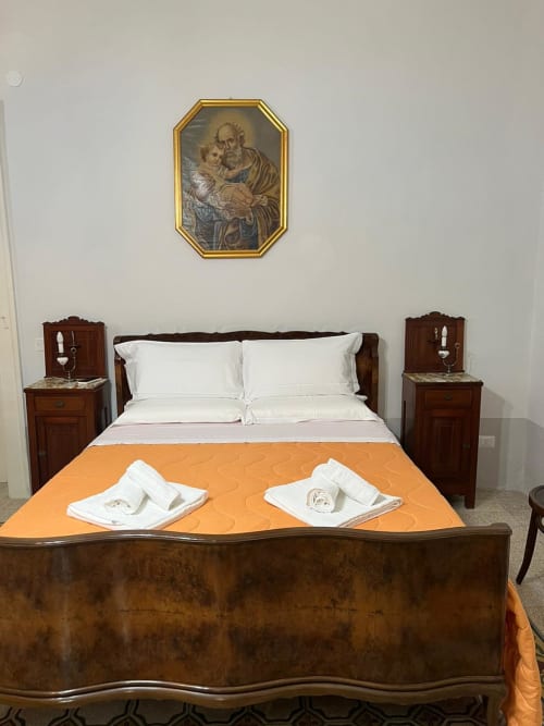 Camera elegante a Palazzo Porta, Procida con letto in legno, biancheria bianca, plaid arancione e opere religiose alle pareti.