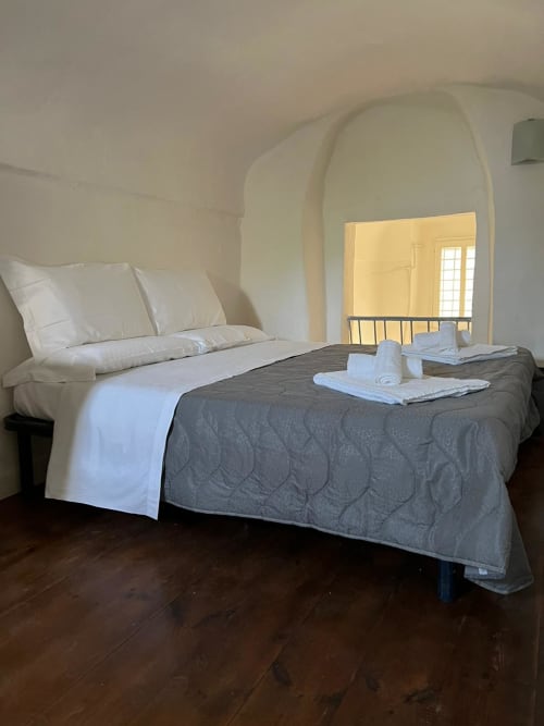 Camera da letto di Palazzo Porta a Procida con letto, cuscini bianchi, coperta grigia e porta ad arco con vista sul giardino