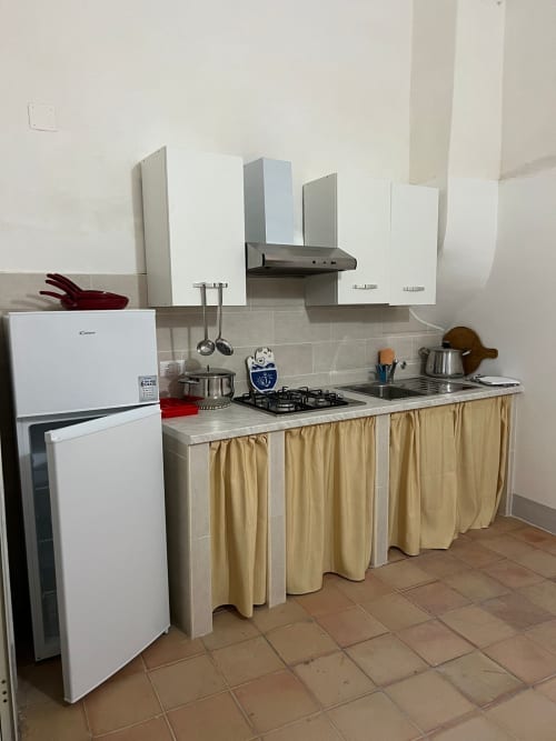 Cucina compatta a Palazzo Porta BnB a Procida con mobili bianchi, fornello a gas, frigorifero e tenda beige per riporre.