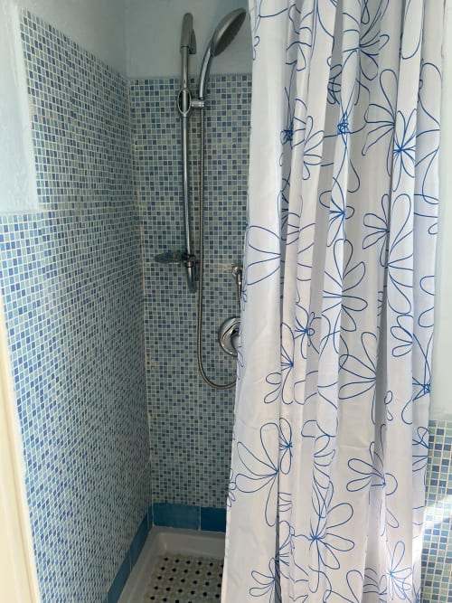 Cabina doccia con pareti rivestite in mosaico azzurro e tenda bianca a fiori presso l'hotel Palazzo Porta, Procida