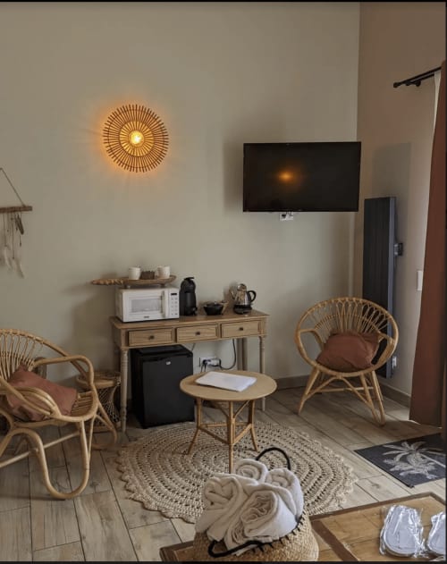 Coin salon douillet à La Cour d'Hortense avec fauteuils en rotin, table ronde, TV murale et luminaire soleil décoratif