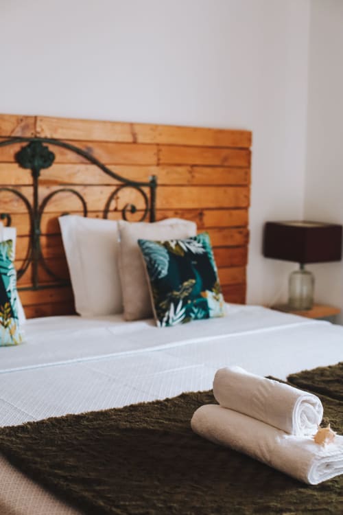 Quarto aconchegante na Kindnessmoon em Óbidos com toalhas enroladas, cabeceira de madeira e almofadas tropicais