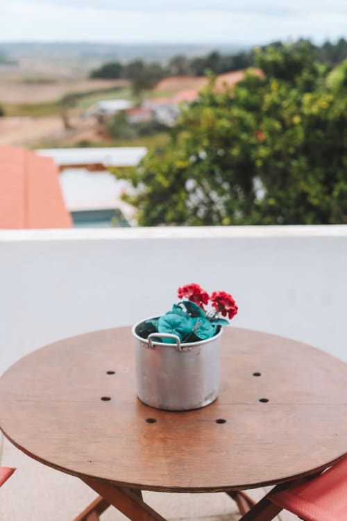 Gerânios vermelhos em vaso branco sobre mesa de madeira na varanda da Kindnessmoon BnB, com vista para a zona rural de Óbidos