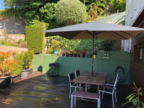 Patio de Casa Arealonga en Vigo con mesa de comedor, sombrilla gris, plantas en macetas y exuberante vegetación