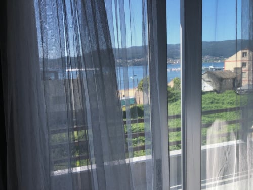 Cortinas translúcidas enmarcan la vista desde el balcón de la bahía de Vigo, colinas y edificios de piedra tradicionales en Casa Arealonga