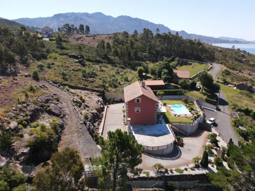 Vista aérea del hotel A Ballenera de Caneliñas en Cee, España, con edificio rojo, piscina y paisaje montañoso costero.