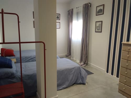 Dormitorio luminoso en Casa Arealonga de Vigo con cama de estructura metálica roja, cortinas a rayas y cómoda de madera