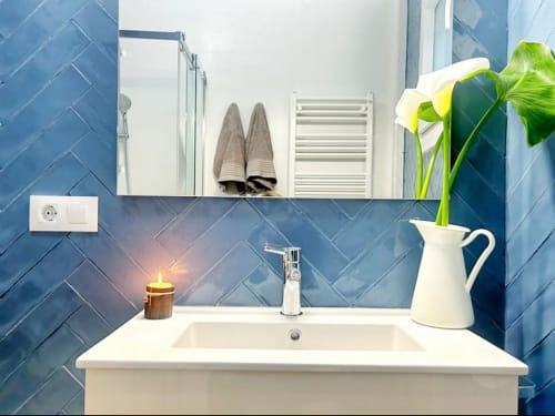 Baño moderno con azulejos de espiga azul, lavabo blanco, espejo y flor amarilla en jarra blanca en Holibai, Baiona