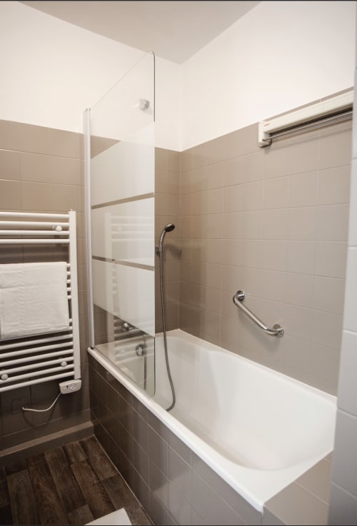 Salle de bain moderne avec baignoire blanche, paroi de douche vitrée, carrelage beige et sèche-serviettes chauffant à Résidence Le Roc Fleuri