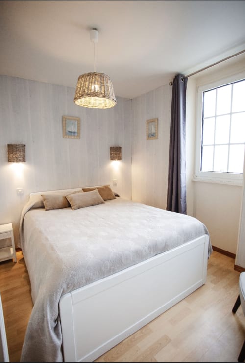Chambre lumineuse à la Résidence Le Roc Fleuri avec lit blanc, luminaire suspendu en rotin et vue sur mer