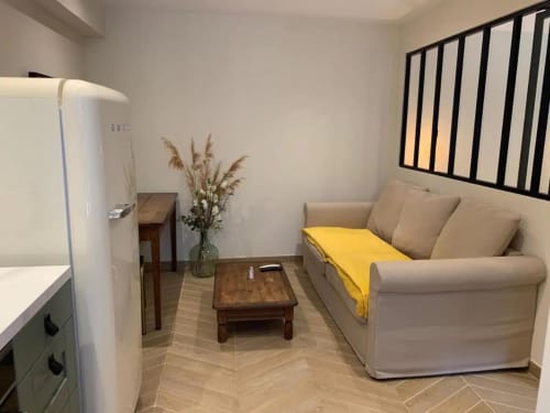 Coin salon confortable à Beaune avec canapé beige, plaid jaune, table basse en bois, réfrigérateur blanc et fenêtre
