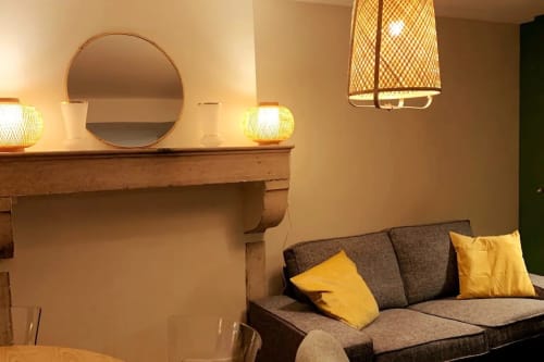 Salon de la villa Beaune avec canapé gris, coussins jaunes, miroir rond, luminaires suspendus en rotin et manteau de cheminée