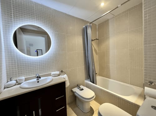 Baño moderno en Apartamentos J&A Terrassa con espejo iluminado, lavamanos blanco, inodoro, bañera y azulejos mosaico beige