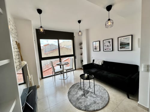 Sala de estar moderna de los Apartamentos J&A en Terrassa con sofá negro, lámparas colgantes y balcón con vistas a la ciudad