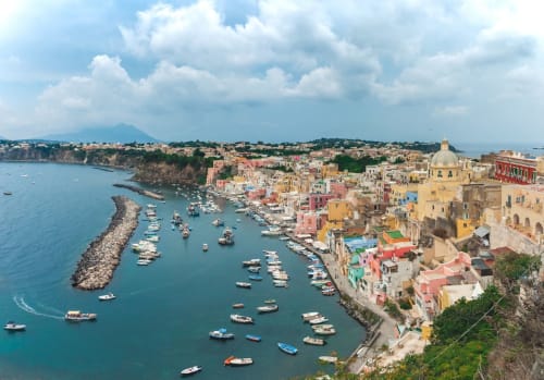 Le colorate case a picco sul mare di Procida si affacciano sul porto turchese con barche ormeggiate e frangiflutti roccioso