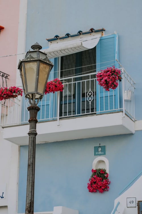 Facciata del Palazzo Porta B&B a Procida con persiane blu, fiori rossi e lampada vintage