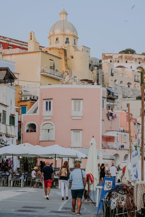 Piazza di Procida con edificio rosa, chiesa dal tetto a cupola, bancarelle di mercato e turisti presso Palazzo Porta