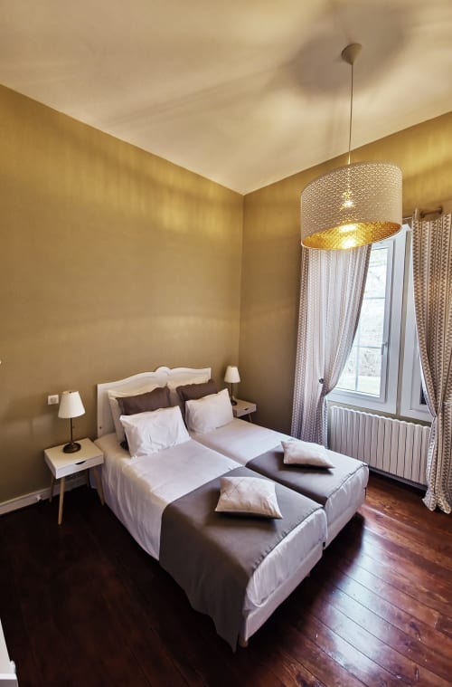 Chambre élégante du Manoir Le Coutureau aux murs dorés, luminaire suspendu, literie blanche et fenêtre aux rideaux à motifs