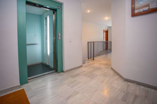 Pasillo moderno de apartamento con ascensor turquesa, pisos de madera y barandillas metálicas en Puerto Santa María
