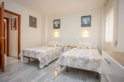 Luminoso dormitorio doble en apartamento de Puerto Santa María con dos camas, cabeceros blancos, ropa de cama con motivos florales y apliques de pared