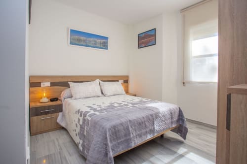 Moderno dormitorio en apartamento Wehost Costaluz, Sanlúcar. Cama de madera, paredes blancas, ventana con vista a la ciudad, lámpara mesita, cuadro