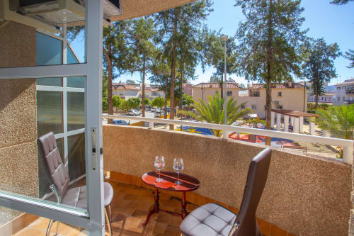 Balcón con vista de la ciudad, dos sillas, mesita con copas de vino, árboles y edificios visibles en Puerto Santa María