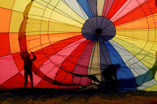 Photographie colorée prise de l'intérieur d'une montgolfière en train d'être gonflée. Le tissu de l'enveloppe présente un motif de rayons de l'arc-en-ciel, avec des panneaux rouges, orange, jaunes, verts, bleus et violets convergeant vers le sommet central. Deux silhouettes de personnes sont visibles à la base, s'affairant autour du matériel de gonflage. Un texte en français est superposé au centre, indiquant : « Gonflage coloré d'une montgolfière dans le Marais Poitevin, France, où se trouve notre maison d'hôtes avec ses 3 chambres