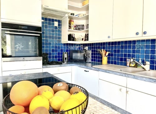 Moderne Küche mit weißen Schränken, blauem Fliesenspritzschutz, Edelstahlspüle und frischem Obst im schwarzen Korb in der Villa