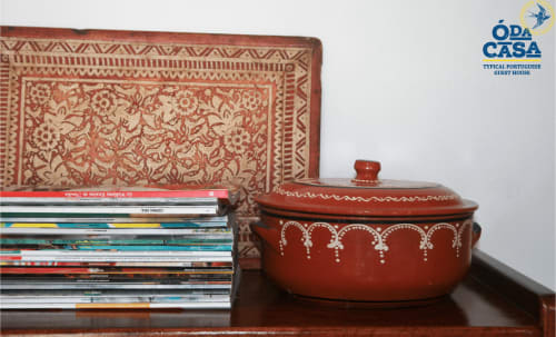 Décor de la maison d'hôtes Ó da Casa : assiette en céramique portugaise et magazines empilés près d'un coussin rouge ornementé à Peniche