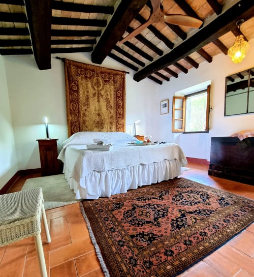 Rustic villa bedroom with exposed wooden beams, white bed, ornate tapestry, terracotta floors, and traditional decor