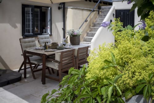 Area patio soleggiata con tavolo da pranzo in legno, sedie imbottite, piante rigogliose e fiori viola