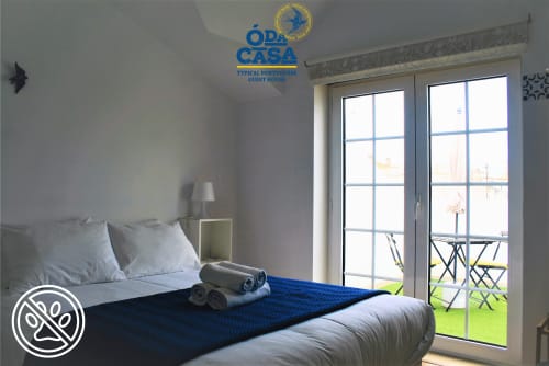 Quarto claro na pensão Ó da Casa em Peniche com acentos azuis, portas de varanda, vista para a cidade e comodidades modernas