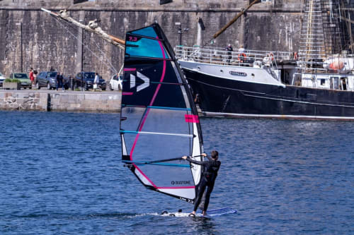 Véliplanchiste en action au large de Belle-Île-en-Mer avec bateaux du port et quai en arrière-plan
