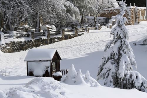 Chalé montanhês coberto de neve em Cota 1500, Penhas da Saúde, com capela, pinheiros e paisagem invernal pristina