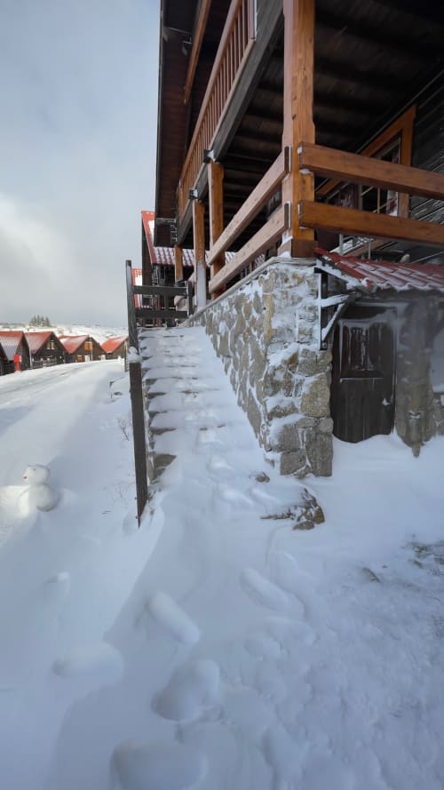 Exterior nevado do chalé Cota 1500 em Penhas da Saúde com deck de madeira e fundação de pedra no inverno