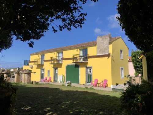 Bright yellow Villa Várzea B&B in Várzea, Portugal with green door, pink chairs, and manicured lawn under blue sky