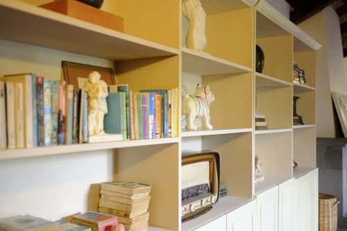 White shelving unit with yellow backing displaying books, decorative sculptures, vintage radio, and woven baskets in Villa
