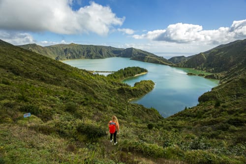 Hiker in red jacket overlooking turquoise crater lake surrounded by lush green mountains near Várzea, Portugal