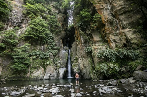 Shirtless man standing in shallow river pool beneath waterfall in lush green canyon, Várzea Portugal