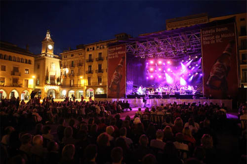 Concert a l'aire lliure nocturn a Vic, Barcelona amb escenari il·luminat en porpra, edificis històrics, torre del rellotge i gran multitud